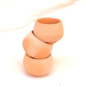 Puede incluir: Tres pequeñas macetas de terracota apiladas, de forma redondeada y con una amplia abertura. Las macetas son de un color marrón anaranjado cálido y terroso, sobre un fondo blanco.