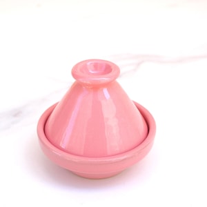 Puede incluir: Una olla de tagine de cerámica rosa, con tapa cónica y base circular. La tapa tiene un pequeño asa redondeada. El tagine es un recipiente de cocina tradicional marroquí, utilizado para guisos y tagines.