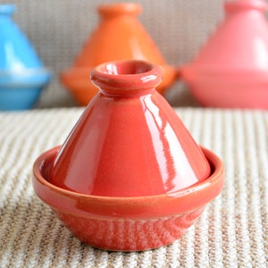 Könnte beinhalten: Ein roter Keramik-Tajine-Topf mit konischem Deckel, auf einer strukturierten beigefarbenen Oberfläche. Weitere Tajine-Töpfe in Blau, Orange und Rosa sind im Hintergrund zu sehen. Dieses traditionelle Kochgeschirr wird für Schmorgerichte verwendet.
