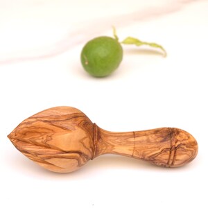 Op de afbeelding: Een houten citruspers met een getextureerde, lichtbruine afwerking. Naast de pers ligt een groene limoen met een steel en bladeren. De pers is ontworpen voor het persen van citrusvruchten.