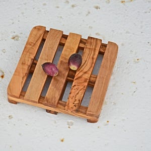 May include: A square wooden coaster with a grid pattern. The coaster is made of light brown wood with a natural finish. Two small purple and brown objects are on the coaster.