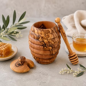 Może przedstawiać: Pojemnik na miód wykonany z drewna oliwnego o wyraźnym naturalnym usłojeniu, posiadający żebrowaną powierzchnię zewnętrzną, dopasowaną okrągłą pokrywkę z małym uchwytem oraz długą drewnianą łyżkę do miodu z rowkowaną końcówką.