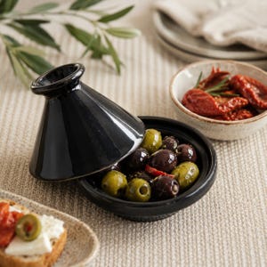 Könnte beinhalten: Eine schwarze Keramik-Tajine mit leicht geöffnetem Deckel, gefüllt mit grünen und dunklen Oliven. Daneben kleine Schalen mit Oliven und sonnengetrockneten Tomaten, für eine mediterrane Vorspeisen-Szene.