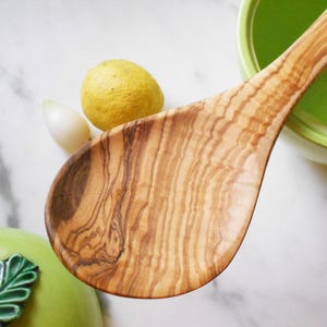 May include: A wooden spoon crafted from olive wood, showcasing a natural wood grain pattern. The spoon is light brown in colour and has a rounded shape.