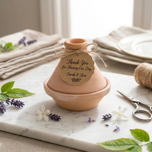 May include: A small, terracotta tagine pot with a lid, tied with twine and a tag that reads "Thank You for Sharing Our Day Sarah & Ben." The pot is surrounded by lavender sprigs and white flowers, suggesting a wedding favor or decorative item.