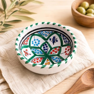 May include: A small, white ceramic bowl with a hand-painted floral design. The bowl features a green, black, blue, and red pattern on the inside and a green dotted border. The bowl is sitting on a beige cloth.