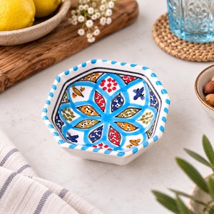 May include: A hexagonal ceramic bowl featuring a white base with a hand-painted blue, red, yellow, and green floral pattern. The rim is decorated with a repeating blue and white dash design, and the interior displays a symmetrical petal motif.