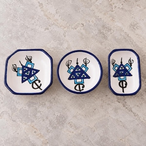 Op de afbeelding: Drie witte keramische schaaltjes met blauwe randen en handgeschilderde geometrische ontwerpen in blauwe en zwarte tinten. De set bevat een achthoekig, een rond en een rechthoekig schaaltje, elk met een centraal abstract driehoekig motief.