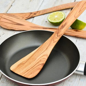 Könnte beinhalten: Drei Holzspatel mit natürlicher Holzmaserung, einer liegt in einer schwarzen Bratpfanne mit schwarzem Griff. Die Spatel sind aus Olivenholz und eignen sich ideal zum Kochen und Servieren.