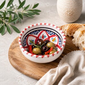 Może przedstawiać: Mała, biała ceramiczna miska z kolorowym wzorem, wypełniona oliwkami, suszonymi pomidorami i ziołami. Miska stoi na drewnianej desce, z chlebem i dekoracyjnym wazonem w tle. Widoczna jest gałązka oliwna.