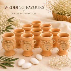 Puede incluir: Pequeños tarros de terracota, ideales para recuerdos de boda, se exhiben. Cada tarro está adornado con un lazo de cuerda y una etiqueta que dice "Thank You". Dulces blancos y ovalados están dispersos. El texto "WEDDING FAVOURS" es visible.