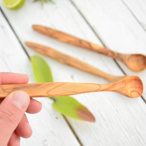 Könnte beinhalten: Drei Holzlöffel mit abgerundeten Schalen. Die Löffel sind aus hellem braunem Olivenholz gefertigt und haben eine natürliche, rustikale Oberfläche.