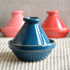 May include: A teal blue ceramic tagine with a lid. The tagine is sitting on a woven rug with other tagines in the background.