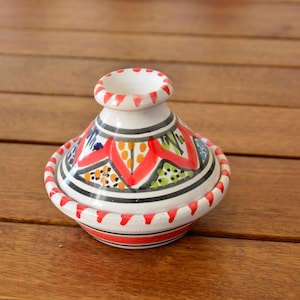 May include: A white ceramic tagine with a red, orange, green and blue geometric pattern. The tagine is on a wooden surface.