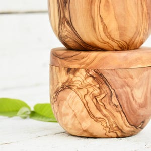 Può includere: Due ciotole in legno d'ulivo impilate. Le ciotole presentano un motivo a venature naturali del legno in tonalità marroni e beige. La forma liscia e arrotondata suggerisce che siano per servire o conservare.
