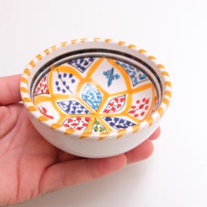 May include: A small, white ceramic bowl with a hand-painted floral design. The interior features a vibrant pattern of yellow, blue, red, and green shapes. The rim is decorated with a yellow and orange dotted pattern, and black and white stripes.