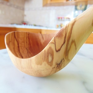 May include: A wooden ladle with a natural wood grain finish. The ladle is made of olive wood and has a deep bowl.
