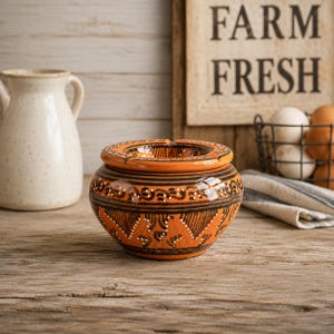 May include: A round, orange ceramic ashtray featuring a glossy finish and intricate black hand-painted patterns. The design includes horizontal bands, swirling motifs, and small white dots encircling the exterior and the wide, flat rim.