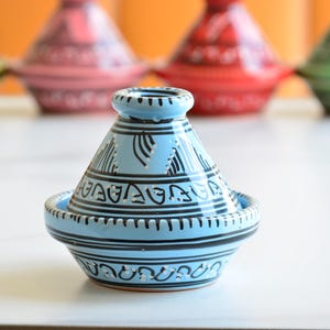 Op de afbeelding: Een kleine, lichtblauwe keramische tajine met zwarte geometrische ontwerpen. De tajine heeft een ronde basis en een conische deksel.