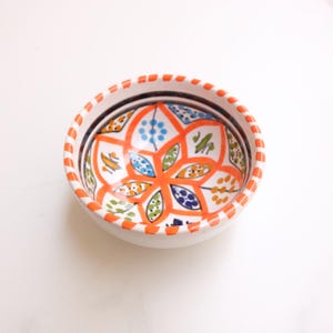 Può includere: Una piccola ciotola in ceramica bianca con un vivace motivo floreale. L'interno presenta un motivo floreale arancione con accenti blu, verdi e gialli. Il bordo è decorato con trattini arancioni. Un pezzo decorativo per servire o esporre.