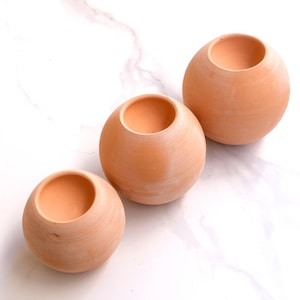 Könnte beinhalten: Drei kugelförmige Pflanzgefäße aus Terrakotta in verschiedenen Größen. Jedes Pflanzgefäß hat eine breite Öffnung und eine glatte, unglasierte Oberfläche. Die Pflanzgefäße sind auf einer weißen Marmoroberfläche angeordnet.
