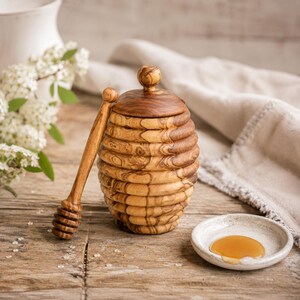 May include: A wooden honey pot with a lid and a honey dipper. The pot is shaped like a beehive and has a natural wood grain finish. A small ceramic dish contains a pool of honey. The background includes a white vase and flowers.