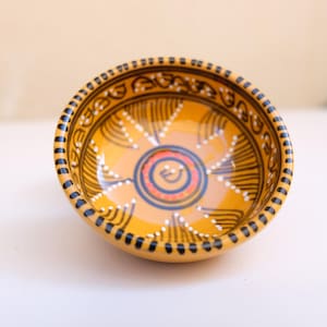Op de afbeelding: Een kleine, ronde keramische kom met een heldergele binnen- en buitenkant. De kom heeft een gedetailleerd handgeschilderd ontwerp met zwarte, witte en blauwe accenten. De rand is versierd met een reeks zwarte stippen. Het ontwerp van de kom is gecentreerd met een spiraal.