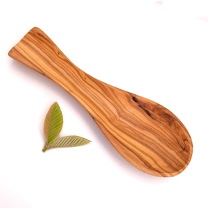 Può includere: Un cucchiaio di legno con una ciotola ampia e curva e un lungo manico. Il cucchiaio è in legno marrone chiaro con un motivo a venature visibile. Due foglie verdi sono accanto al cucchiaio su sfondo bianco. Un utensile da cucina.