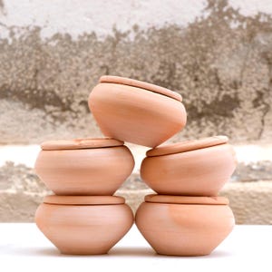 Op de afbeelding: Een stapel van zes kleine, ronde potten in terracotta kleur met deksels. De potten zijn gerangschikt in een piramidevorm, waarbij de deksels elkaar lichtjes overlappen. De achtergrond is een getextureerde, neutraal getinte muur.