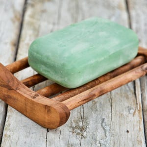 Könnte beinhalten: Eine grüne Seife liegt auf einem hölzernen Seifenschälchen. Die Schale hat eine geschwungene Form und ist aus Naturholz gefertigt.