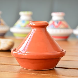 Può includere: Un piccolo tagine di terracotta color terracotta con un coperchio arrotondato. Il tagine è appoggiato su una superficie di legno con altri tagine sullo sfondo.