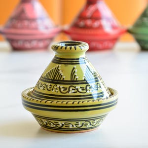 May include: A yellow ceramic tagine with black and white geometric designs. The tagine is sitting on a white surface.