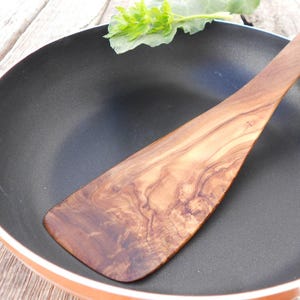 Op de afbeelding: Een houten spatel met een natuurlijk houtnerfpatroon, liggend in een zwarte koekenpan.