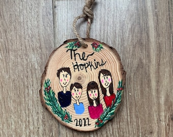 May include: A wooden Christmas ornament with a painted family portrait and the text "The Hopkins 2022". The ornament is decorated with green holly leaves and red berries.