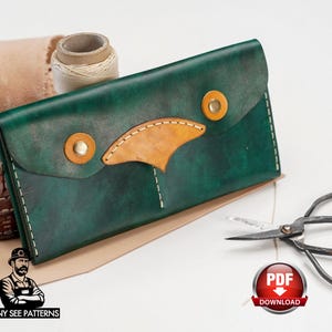 May include: A handcrafted, dark green leather wallet with a tan accent and gold-colored rivets. The wallet is rectangular with a flap closure. A spool of thread, scissors, and a PDF download badge are also visible.