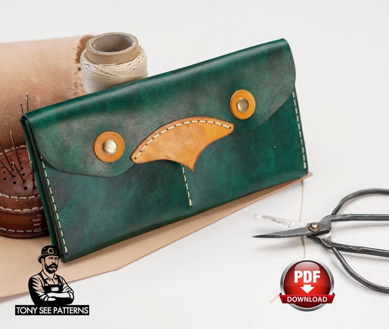 May include: A handcrafted, dark green leather wallet with a tan accent and gold-colored rivets. The wallet is rectangular with a flap closure. A spool of thread, scissors, and a PDF download badge are also visible.