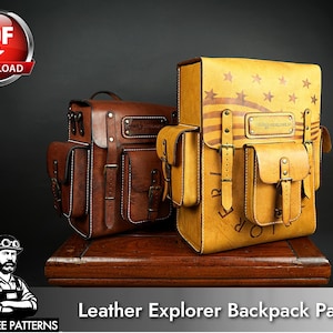 May include: Two leather backpacks with side pockets and front straps. One is dark brown, and the other is tan with a printed American flag design and the text "EXPLORER" visible on the lower front. Both feature metal buckles and rivets.