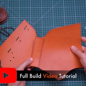 May include: A tan leather piece with pre-punched stitching holes and slots, featuring a central spine section. The leather is unfolded and flat, showing a smooth surface texture. A black banner with the text Full Build Video Tutorial is visible.