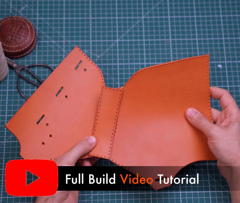 May include: A tan leather piece with pre-punched stitching holes and slots, featuring a central spine section. The leather is unfolded and flat, showing a smooth surface texture. A black banner with the text Full Build Video Tutorial is visible.