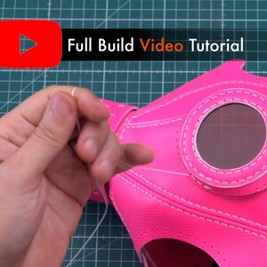 May include: A close-up of a pink leather mask with a white stitch detail. The mask has a round opening for a lens and is being sewn together with white thread.