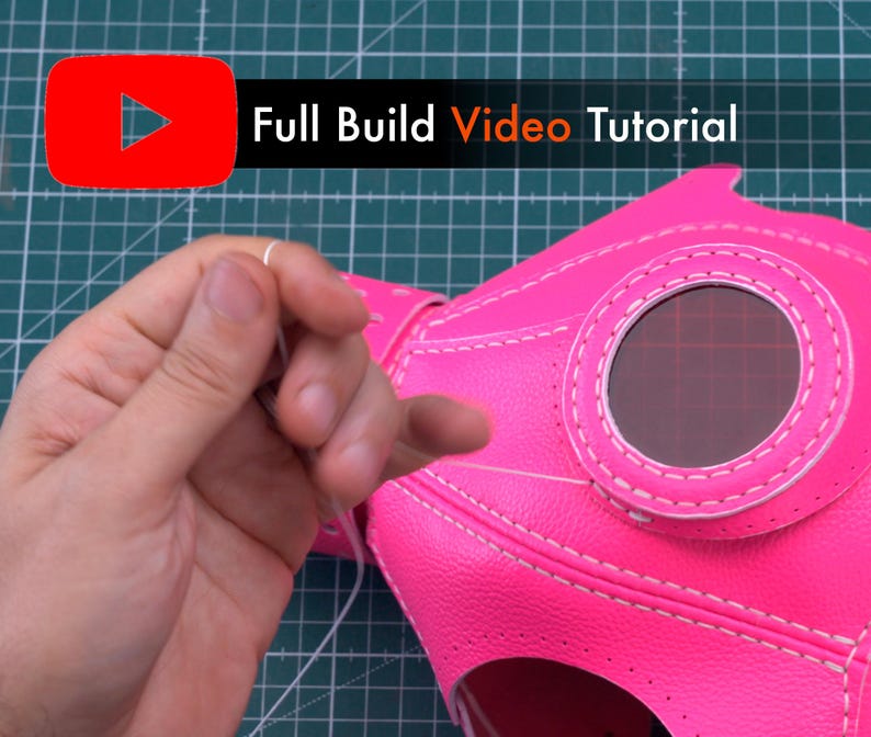 May include: A close-up of a pink leather mask with a white stitch detail. The mask has a round opening for a lens and is being sewn together with white thread.