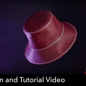 Puede incluir: Un sombrero de pescador color burdeos intenso con un acabado liso similar al cuero y costuras blancas prominentes a lo largo de la copa y el ala. El texto "PDF DOWNLOAD" y "Pattern and Tutorial Video" aparece en la parte inferior.