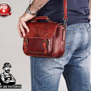 May include: A reddish-brown leather satchel with a top handle and adjustable shoulder strap. The bag features two front buckles and white stitching. The bag is being carried by a person wearing blue jeans.