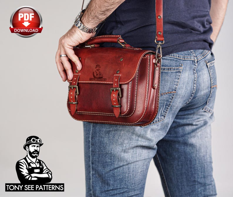 May include: A reddish-brown leather satchel with a top handle and adjustable shoulder strap. The bag features two front buckles and white stitching. The bag is being carried by a person wearing blue jeans.