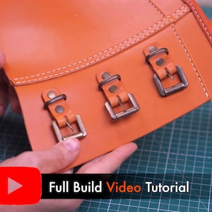 May include: A smooth orange leather panel featuring three metallic buckles secured with rivets and white stitching. The leather piece has a rectangular shape with a reinforced edge. A red YouTube icon and the text Full Build Video Tutorial appear.