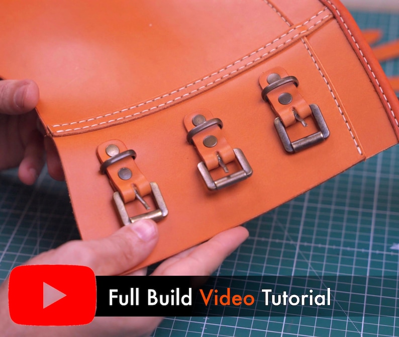 May include: A smooth orange leather panel featuring three metallic buckles secured with rivets and white stitching. The leather piece has a rectangular shape with a reinforced edge. A red YouTube icon and the text Full Build Video Tutorial appear.