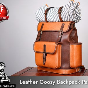Puede incluir: Una mochila de cuero bicolor en marrón y beige con un bolsillo frontal, bolsillos laterales y correas con hebillas ajustables. Una manta enrollada con estampado blanco y negro está sujeta a la parte superior con correas de cuero y hebillas.