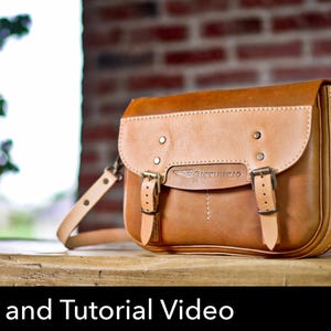 Könnte beinhalten: Eine hellbraune Ledertasche mit Klappverschluss, zwei vorderen Schnallenriemen und Metallbeschlägen. Die Tasche zeigt ein eingeprägtes Logo mit einem Flügelsymbol und dem Text "LEATHERCRAFT" auf der Vorderseite. Sie hat einen verstellbaren Schulterriemen.