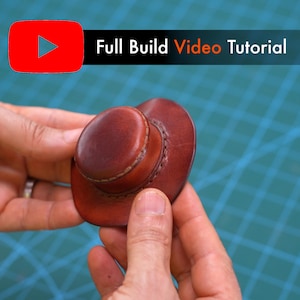 May include: A miniature brown leather cowboy hat featuring visible stitching around the crown and brim. The text "Full Build Video Tutorial" is displayed in white and orange lettering next to a red YouTube play icon above the object.