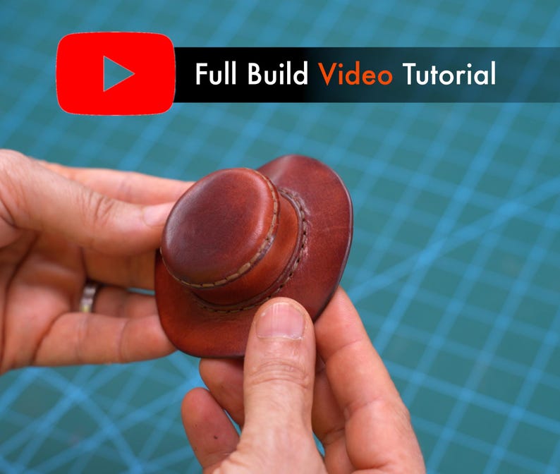 May include: A miniature brown leather cowboy hat featuring visible stitching around the crown and brim. The text "Full Build Video Tutorial" is displayed in white and orange lettering next to a red YouTube play icon above the object.